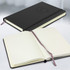 A black hardcover notebook with a branded logo, featuring lined pages and a ribbon bookmark, open and closed.