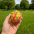 A hand holds a brightly coloured, textured rubber ball with swirls of pink, yellow, and blue against a green park backdrop.