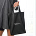 A black tote bag with two handles, featuring a logo on one side, held by a person.
