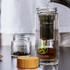 A drink bottle made of clear glass with a metal infuser, wooden lid, and a logo. Contains brewing tea.