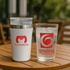 A drink bottle and glass, one white with a silver band and logo, the other clear with a logo, both on a wooden table.