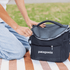 A black cooler bag with a zippered top, featuring a handle and a logo, placed on a picnic blanket.