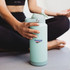 A drink bottle in mint green with a straw lid and a logo, held by a person sitting on a mat.