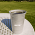 A recycled aluminum stadium party cup in silver, filled with a dark beverage, set on a table in a grassy area.