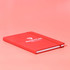 A red A5 notebook with a logo on the cover and an elastic band closure, set against a pink background.
