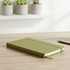 A green notebook with a textured cover and elastic band, placed on a wooden surface near potted plants.