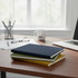 A stack of notebooks in navy, green, and yellow on a wooden table beside a laptop and a coffee mug.