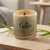 A bamboo candle with a wooden wick, featuring a logo, set on a wooden table with green foliage.
