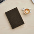 A dark brown notebook with a textured cover rests on a light wooden surface next to a cup of latte art.