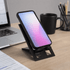 A black foldable phone stand with a phone displaying a gradient screen, set on a desk with a laptop and mug.