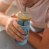 A light blue glass tumbler with a bamboo lid and stainless steel straw, held by a person. It features a logo.