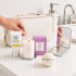 A cream toiletry bag with a candle, bath bomb, hand cream, and a compact mirror, arranged on a bathroom countertop.
