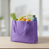A purple non-woven tote filled with bananas, oranges, asparagus, and a drink bottle. It has two handles.