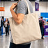 A person holds a beige 450gsm Victorian canvas tote bag with shoulder straps. The bag features a logo.
