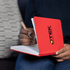 A red A5 notebook with a logo on the cover, held open with a hand writing inside.