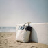 A beach tote bag in cream and navy rests against a white wall on sandy shore, with the ocean in the background.