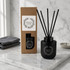 A black botanical reed diffuser with essential jasmine fragrance, accompanied by a brown box and black sticks, on a marble surface.