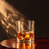 A rocks glass filled with amber liquid and ice cubes, placed on a wooden surface with soft shadows.