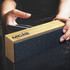 A Bluetooth speaker featuring a bamboo design and a dark fabric front, being held near a smartphone.