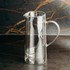A drink jug made of clear glass with a curved handle and spout, featuring a sleek, modern design.