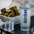 A shot glass featuring the logo "The BOTTLERY," accompanied by a bowl of olives and a cube of ice on a slate surface.