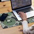 A desk mat featuring a leaf pattern in green and white, with a logo design, beside a laptop and smartphone.