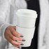 A white ceramic coffee mug with a removable silicone sleeve and lid, held in a hand.