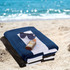 A stack of beach towels in navy blue and black, with sunglasses and sunscreen, placed on sandy beach.