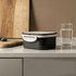 A black and white plastic lunch box on a kitchen countertop, beside a glass of water and a wooden cutting board.