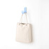 A cream-coloured tote bag hanging on a blue hook against a white wall. The bag has a simple design and a logo.