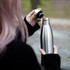 A drink bottle with a sleek stainless steel finish and a black lid, being held by a person outdoors.