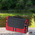 A red and black checked picnic rug with a black handle, resting on a wooden surface near water.