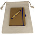 A drawstring bag in cream holds a brown JournalBook with a blue elastic band and a blue pen on top.