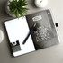 A black and white Nova Bound JournalBook with blank pages, a pen, and a decorative plant on a grey surface.