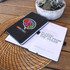 A black notebook with a colourful gear design, open to display its pages. It features a branding logo and a textured cover.