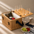 A wicker picnic basket in light brown with a bamboo table, containing wine bottles, glasses, and fresh fruit.