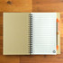 A tan notebook with lined pages and a matching orange pen. The notebook features a spiral binding and a logo.
