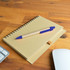 A brown spiral notebook with a blue pen resting on top, placed on a wooden surface alongside a keyboard and mouse.