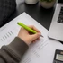 A hand holding a green plastic pen over printed documents and a laptop, with a plant in the background.