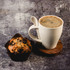 A white wheat fibre cup with coffee, accompanied by a chocolate chip muffin on a wooden coaster.