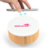 A wireless speaker and charger with a wooden base, featuring a white top and a logo. A hand is holding a smartphone above it.