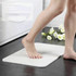 A soft white bath mat with a foot stepping onto it, placed on a tiled floor near flowers. It has a logo.