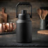 A drink bottle in matte black with a sturdy handle, set against a rustic kitchen backdrop.