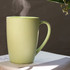A reusable bamboo mug in a soft green colour, emitting steam, sits on a book with foliage in the background.