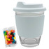 A clear Carlo glass coffee cup with a light blue silicone lid and sleeve, alongside a pack of colourful jelly beans.
