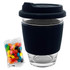 A clear glass coffee cup with a black silicone sleeve and lid, accompanied by a packet of colourful jelly beans.