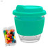 A teal glass coffee cup with a silicone sleeve and lid, accompanied by a pack of multi-coloured jelly beans.