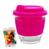 A reusable glass coffee cup with a bright pink silicone sleeve and lid, alongside a pack of colourful jelly beans.