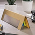 A ruler-shaped sticky note pad in yellow, blue, and green, presented in a brown cardboard holder with a logo.