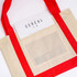A beige reusable grocery bag with red handles features a clear pocket, holding a magazine inside.
